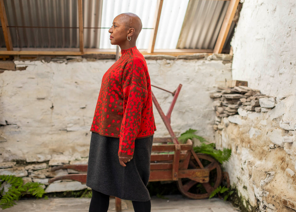 Jeanette Sloan, a black woman with a shaved head stands inside a rustic, whitewashed, stone building in Hoswick Shetland. She is wearing largish, silver hooped earrings with a bead hanging, a tomato red and chestnut brown finely knitted boxy jumper with a graphic animal print patter and a high neck, a carcoal grey woolen dress and black opaque tights. The shed has a corraguated tin rood with skylight inserts. In the background, some ferns grow around an old wooden wheelbarrow painted rusty red.
