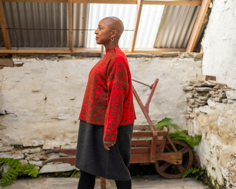 Jeanette Sloan, a black woman with a shaved head stands inside a rustic, whitewashed, stone building in Hoswick Shetland. She is wearing largish, silver hooped earrings with a bead hanging, a tomato red and chestnut brown finely knitted boxy jumper with a graphic animal print patter and a high neck, a carcoal grey woolen dress and black opaque tights. The shed has a corraguated tin rood with skylight inserts. In the background, some ferns grow around an old wooden wheelbarrow painted rusty red.