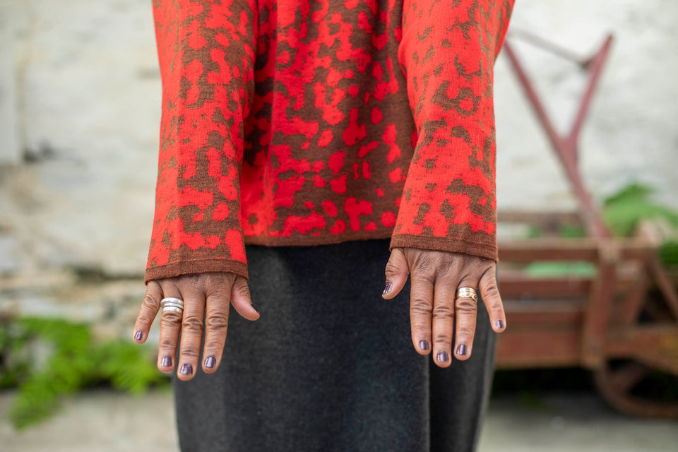 Detail of finely knitted jumper in a red and brown animal print pattern. The model is a black woman, with just her hands seen. She wears chunky silver rings and has her nails painted aubergine. She's also wearing a charcoal grey woollen dress. In the background is an old, wooden wheelbarrow painted rust red. Ferns grow in the cracks between stone floor and whitewashed wall.