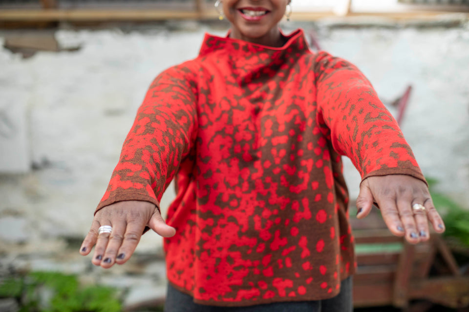 Detail of finely knitted jumper in a red and brown animal print pattern. The model is a black woman, with just the lower part of her face and her hands seen. She smiles and wears chunky hooped silver earrings and chunky silver rings and has her nails painted aubergine. She's also wearing a charcoal grey woollen dress. In the background is an old, wooden wheelbarrow painted rust red. Ferns grow in the cracks between stone floor and whitewashed wall.