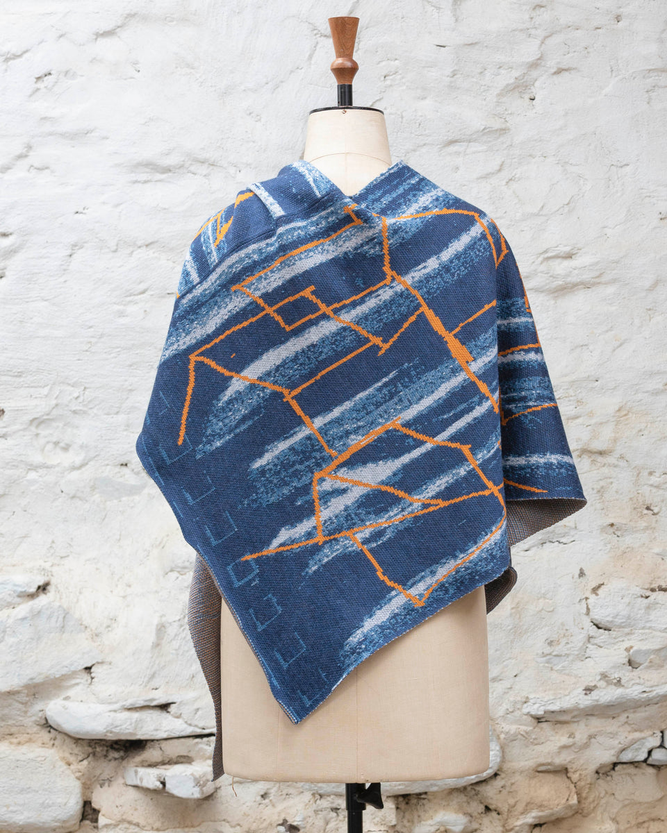 A blue poncho with orange linear design, a statement piece of contemporary knitwear by Nielanell, Shetland, displayed on a mannequin to show pattern