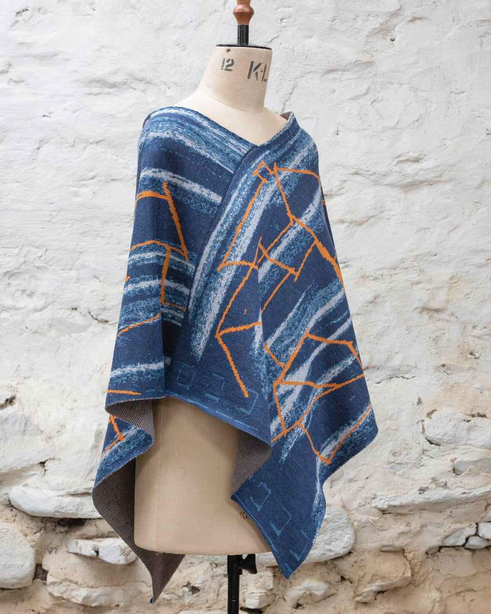 A blue poncho with orange linear design, a statement piece of contemporary knitwear by Nielanell, Shetland, displayed on a mannequin in a rustic stone building in Shetland