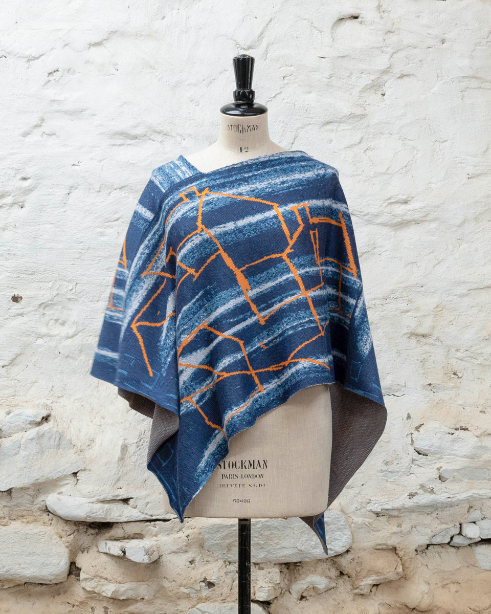 A blue poncho with orange linear design, a statement piece of contemporary knitwear by Nielanell, Shetland, displayed on a mannequin in a stone bothy, Shetland Islands