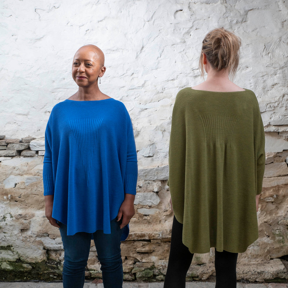 A black woman stands facing the camera and looking off to the side. She is wearing a bright royal blue jumper in a loose style with nipped in sleeves over dark jeans and lace up brown boots. On her left, with her back to the camera, a white woman wears the same jumper in mossy green over black leggings and brown dealer boots.
