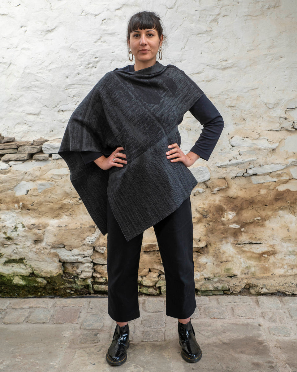 A woman with olive skin and dark hair tied back wears a mid length poncho in a mid grey and charcoal linear design. The shape is asymmetric with different openings for arms on either side. Finely knitted, contemporary Scottish knitwear