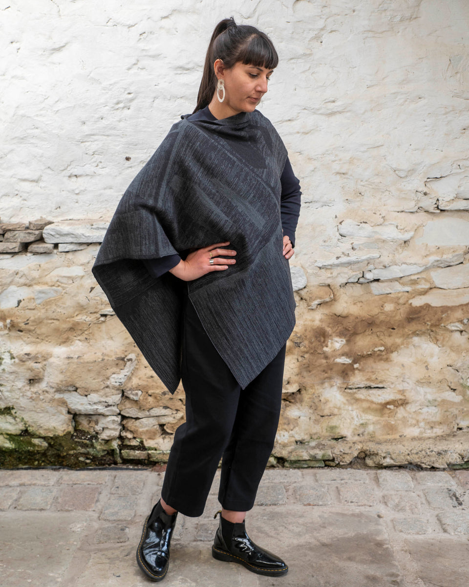 A woman with olive skin and dark hair tied back wears a mid length poncho in a mid grey and charcoal linear design. The shape is asymmetric with different openings for arms on either side. Finely knitted, contemporary Scottish knitwear