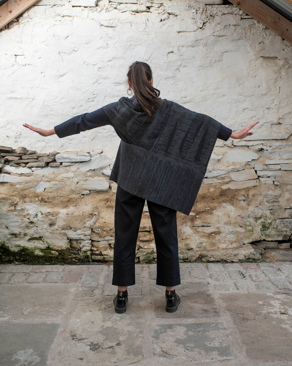 A woman with olive skin and dark hair tied back wears a mid length poncho in a mid grey and charcoal linear design. The shape is asymmetric with different openings for arms on either side. Finely knitted, contemporary Scottish knitwear