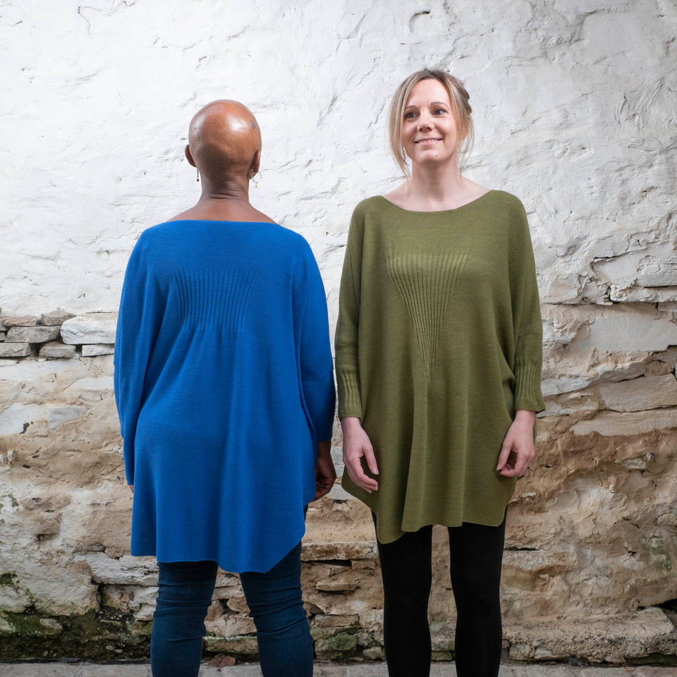 A black woman stands with her back to the camera. She is wearing a bright royal blue jumper in a loose style with nipped in sleeves over dark jeans and lace up brown boots. On her left, facing the camera, a white woman wears the same jumper in mossy green over black leggings and brown dealer boots.
