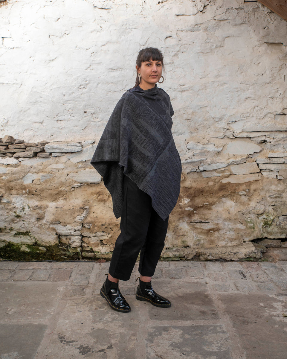 A woman with olive skin and dark hair tied back wears a mid length poncho in a mid grey and charcoal linear design. The shape is asymmetric with different openings for arms on either side. Finely knitted, contemporary Scottish knitwear