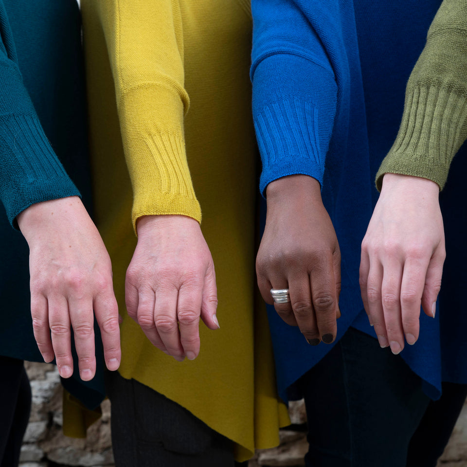 Three white women and one black woman hold their arms out to show the cuffs of their lightweight jumpers. Knitted in dark teal, yellow, bright royal blue and mossy green.