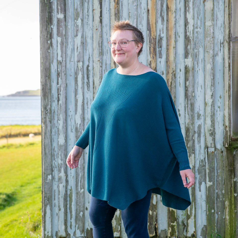 A white woman with short fair hair walks in front of the Hoswick Gospel Hall, Shetland. It's wooden chapel that is now worn by weather. She is wearing clear framed spectacles and an airy dark teal, poncho sweater . The body of the jumper is full and loose and it has dolman sleeves. The jumper swings out as she moves. Underneath she has navy leggings. She turns her head slightly and looks towards the camera.