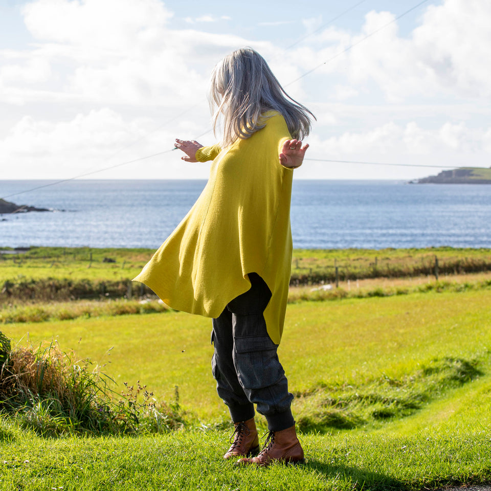 A white woman with long, straight grey hair stands at the top of a field in Shetland overlooking sea. She stands side on to the camera and spins with her arms out. She wears a lightweight, long yellow jumper over charcoal combat trousers and brown lace up boots