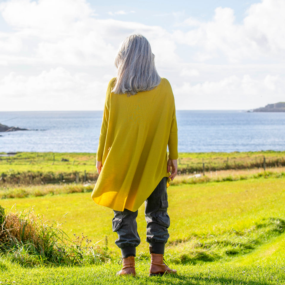 A white woman with long, straight grey hair stands at the top of a field in Shetland overlooking the sea. She wears a lightweight, long yellow jumper over charcoal combat trousers and brown lace up boots