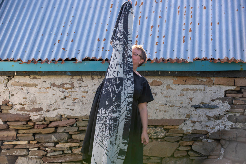 A white woman with short fair hair stands in front of an old stone building with a corrugated tin roof in Hoswick, Shetland. She wears glasses with clear frames and a short sleeved black heavy linen dress with full skirt. She holds up a large shawl/wrap in a contemporary design in charcoal and off white abstract design. Photograph taken in Hoswick, Shetland, Scotland.