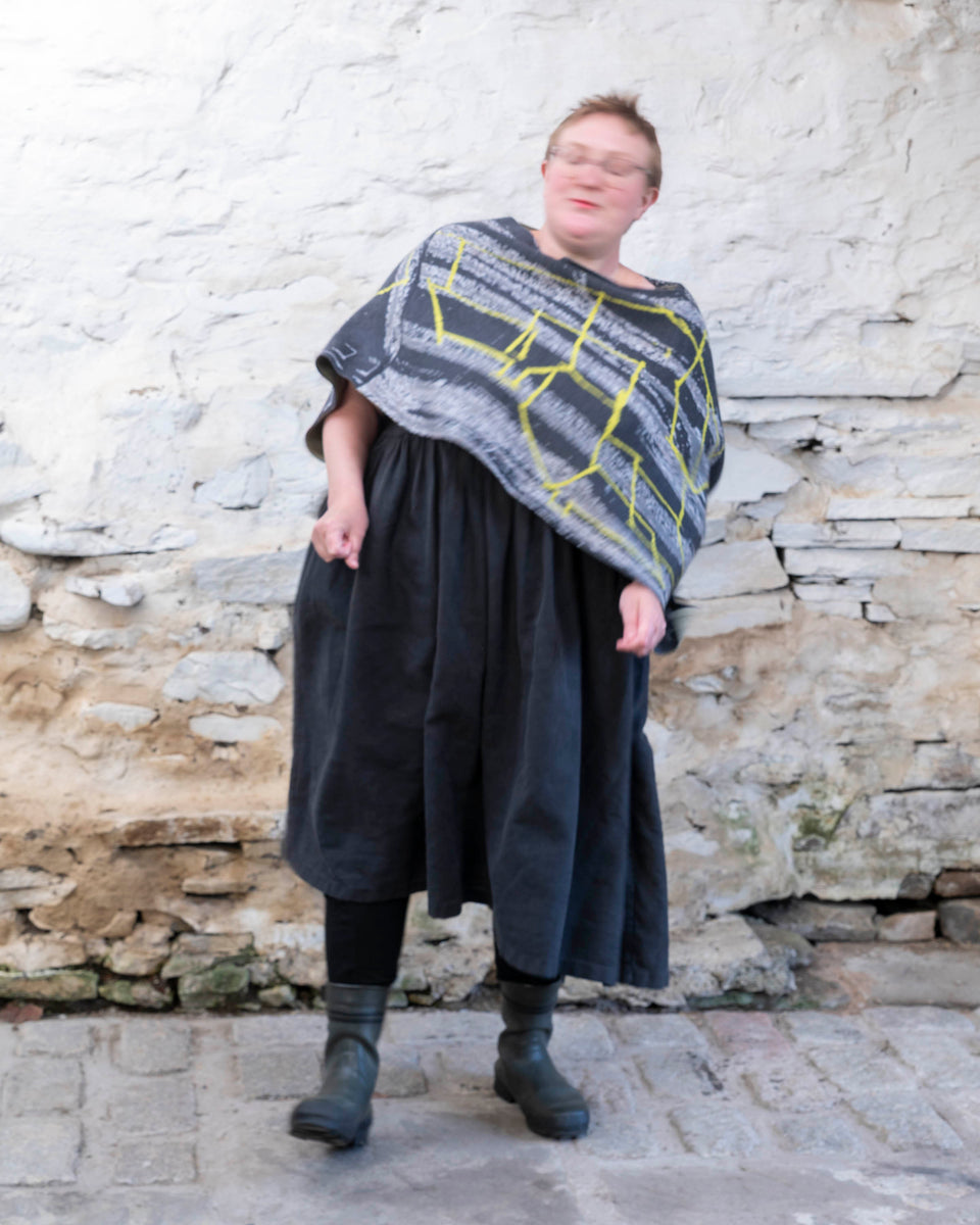 A woman wears a poncho in greys with neon yellow, layered over a black linen dress. Photographed in a stone bothy, Shetland Islands, Scotland.