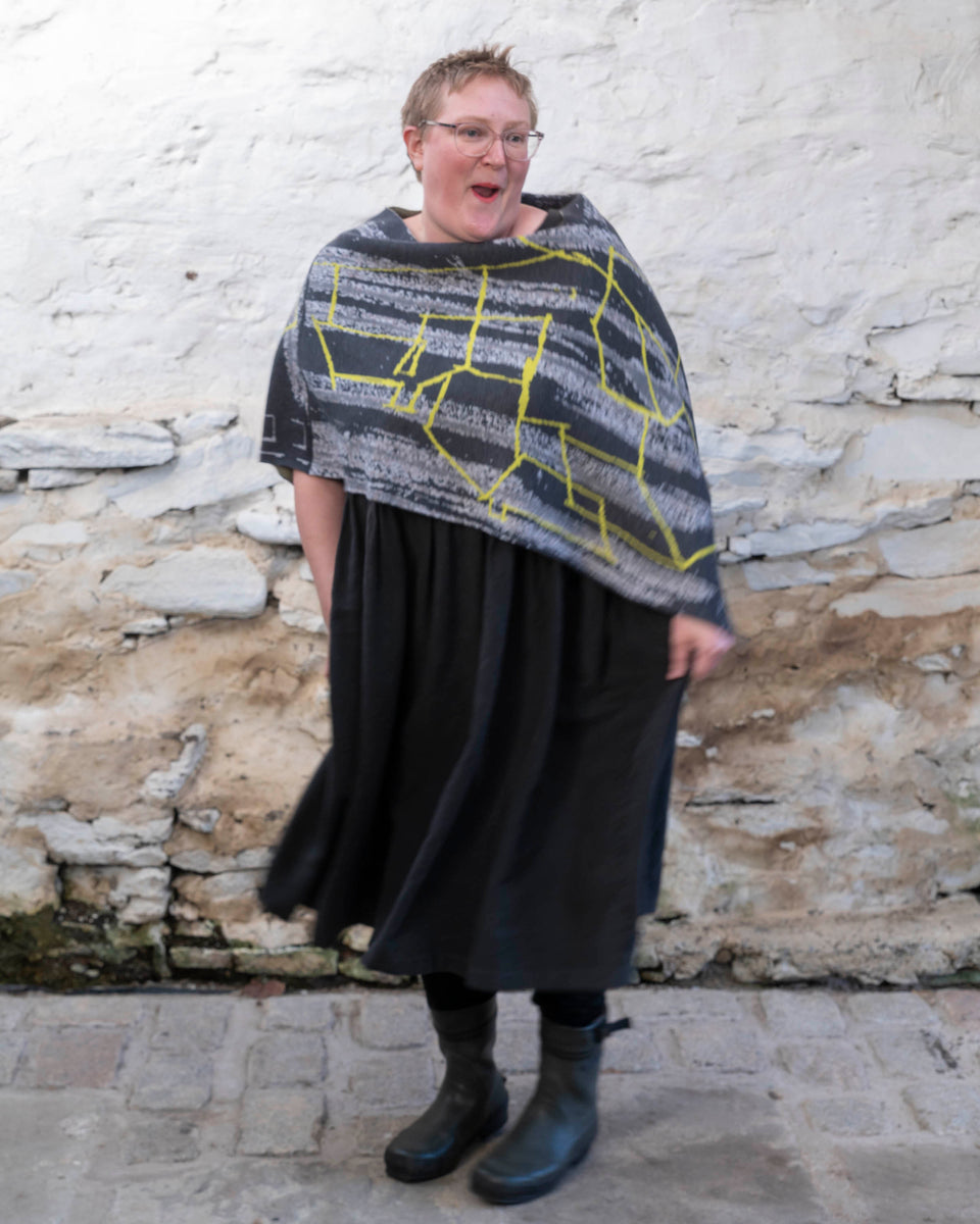 A woman wears a merino poncho in greys with a yellow linear pattern. A statement piece of knitwear.