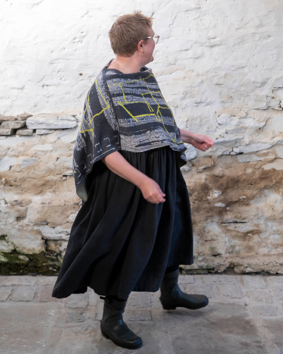 A woman wears a contemporary poncho, a piece of unusual knitwear for layering. In greys with neon yellow linear pattern