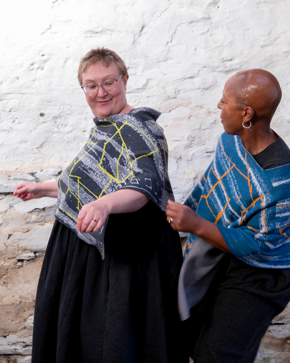 Two women wear capes in a contemporary design one in greys with neon yellow, one with blues and orange. They are layered over charcoal and black dresses.