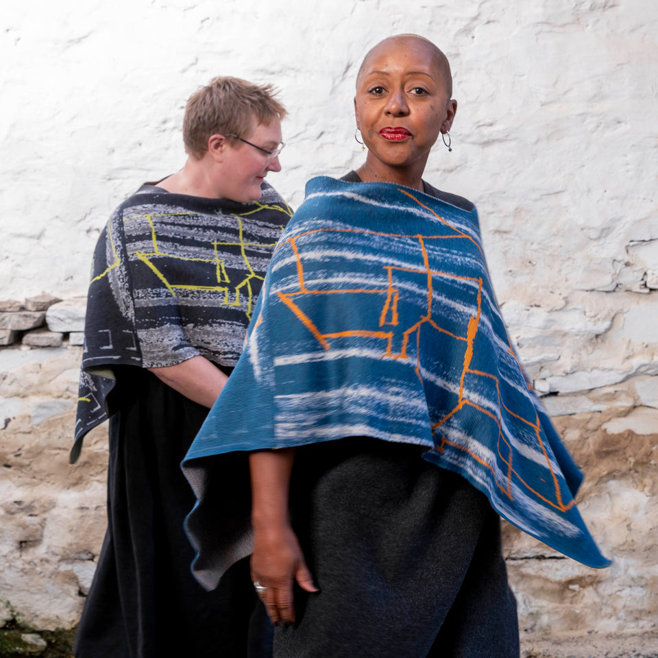 Two women in layered capes in blues and orange and in greys and yellow. Photographed in a rustic stone building, Shetland