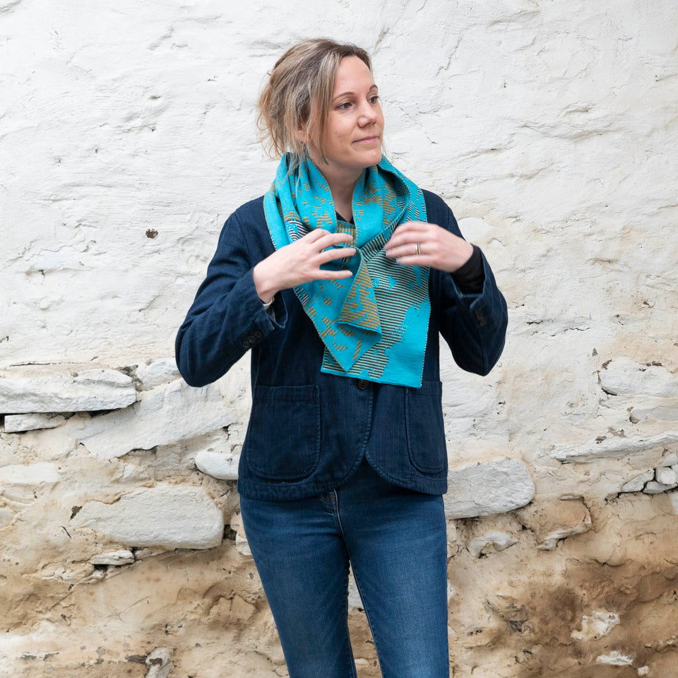 A white woman with fair hair tied back wears a finely knitted scarf made in Shetland, Scotland. It is striped and mottled in interchanging turquoises and mustard yellows. She arranges it arund her neck and looks away from the camera. She is also wearing jeans and a denim jacket.