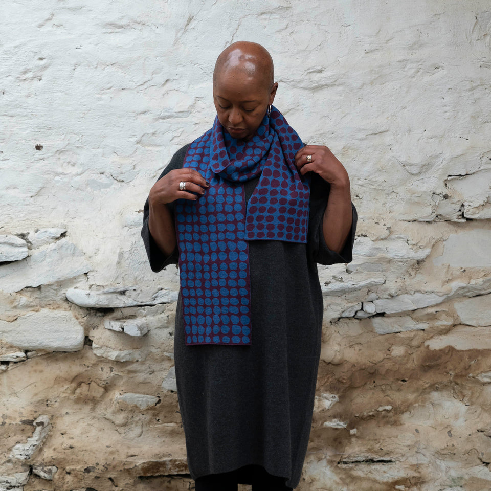 A black woman with a shaved head stands in a rustic whitewashed stone building. She is wearing a finely knitted Scottish scarf in an irregular dot pattern of aubergine and bright blue. She also wears a charcoal wool dress with three quarter length sleeves.