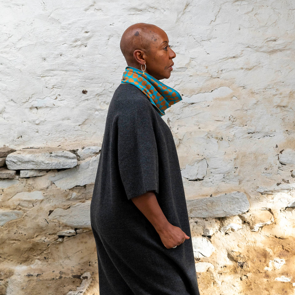 Merino circle scarf - a black woman with shaved head stands in a rustic stone building in Hoswick, shetland. She is wearing a charcoal wool dress with elbow length sleeves . Around her neck is a merino loop scarf in ochre and turquoise.