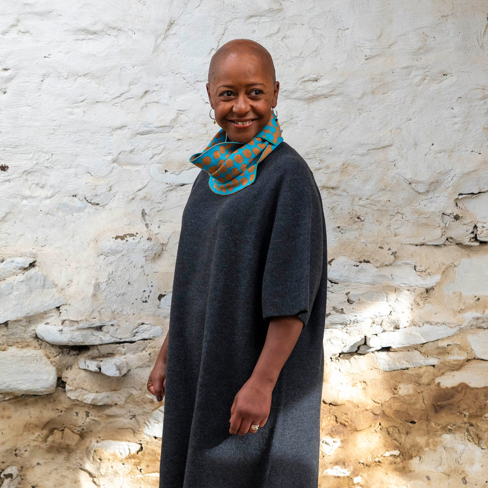 Merino circle scarf - a black woman with shaved head stands in a rustic stone building in Hoswick, shetland. She is wearing a charcoal wool dress with elbow length sleeves . Around her neck is a merino loop scarf in ochre and turquoise.