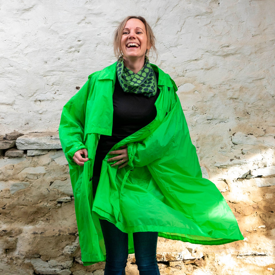Merino loop scarf - A white woman with fair hair tied back stands in a rustic stone building in Hoswick, Shetland Islands, Scotland. She is wearing a green light weight rain coat over jeans and a circle scarf with irregular dot design in green and navy.