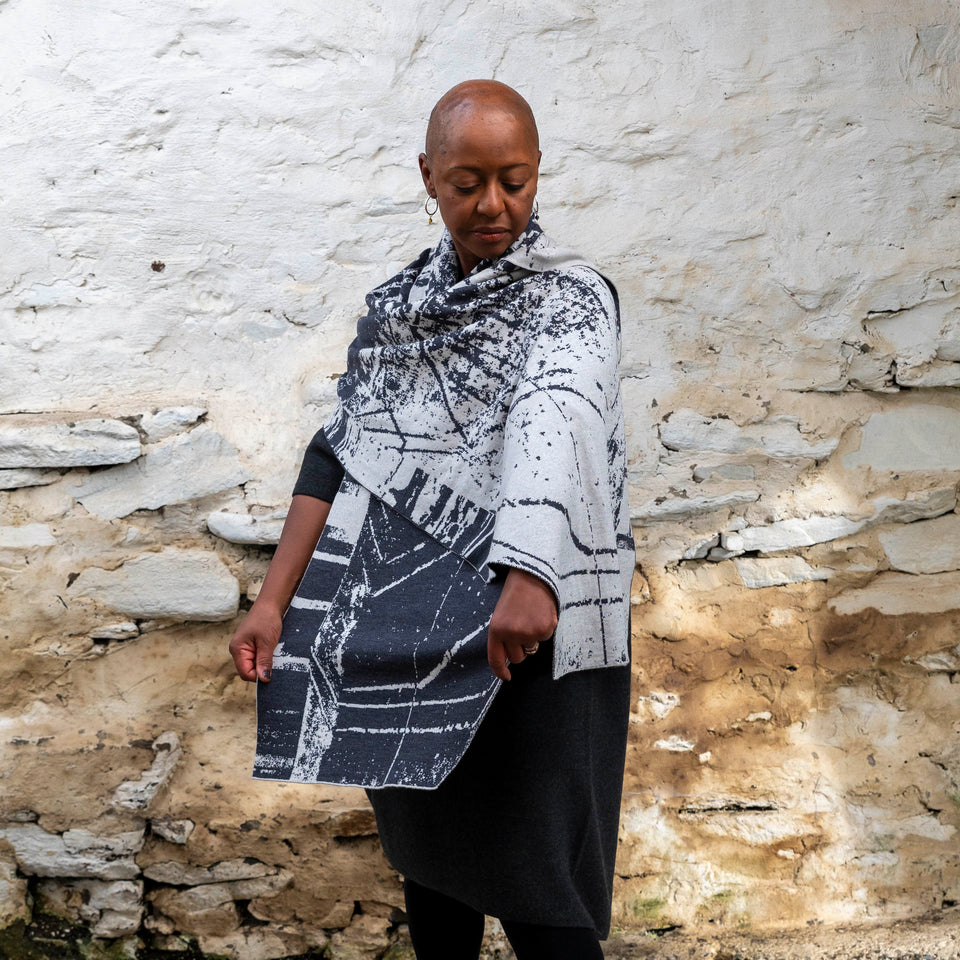 A black woman with a shaved head stands in a rustic stone building with whitewashed walls in Hoswick, Shetland. She wears black tights, a black charcoal woollen dress and has a large charcoal and off-white shawl wrapped around her. She holds out one end.