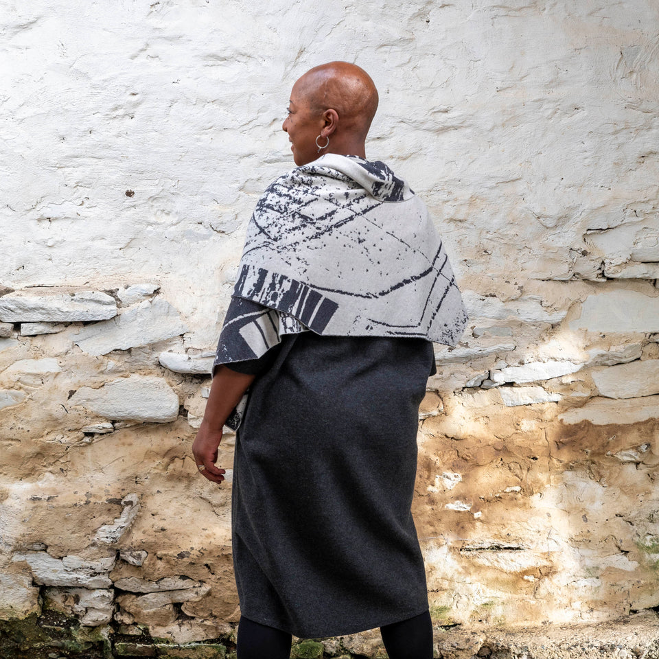 A black woman with a shaved head stands in a rustic stone building with whitewashed walls in Hoswick, Shetland. She wears black tights, a charcoal and off white shawl is wrapped around her. She has her back to the camera showing the angular edges of the shawl