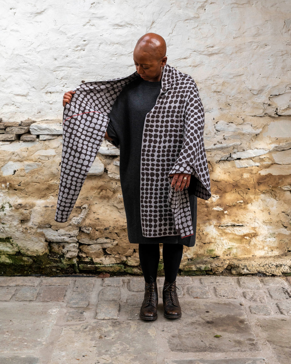 A black woman with a shaved head stands in a rustic stone building with whitewashed walls and a stone flat floor in Shetland.  She wears a finely knitted modern shawl drapaed unfolded around her shoulders and down over her dress.. It is charcoal grey with pale grey irregular dots and a thin meandering line in cherry red. She also wears a charcoal woollen dress in a loose fit, black tights and dark brown brogue boots.