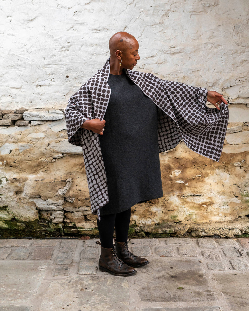 A black woman with a shaved head stands in a rustic stone building with whitewashed walls and a stone flat floor in Shetland. She wears a finely knitted modern shawl drapaed unfolded around her shoulders and down over her dress.. It is charcoal grey with pale grey irregular dots and a thin meandering line in cherry red. She also wears a charcoal woollen dress in a loose fit, black tights and dark brown brogue boots. she stretches her left arm out to show the size of the shawl