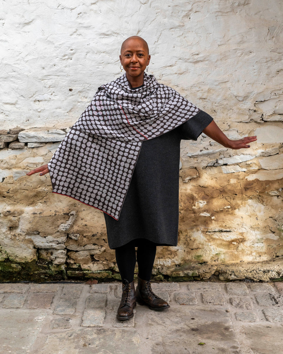 A black woman with a shaved head stands in a rustic stone building with whitewashed walls and a stone flat floor in Shetland. She wears a finely knitted modern shawl drapaed unfolded around her shoulders and down over her dress.. It is charcoal grey with pale grey irregular dots and a thin meandering line in cherry red. She also wears a charcoal woollen dress in a loose fit, black tights and dark brown brogue boots.