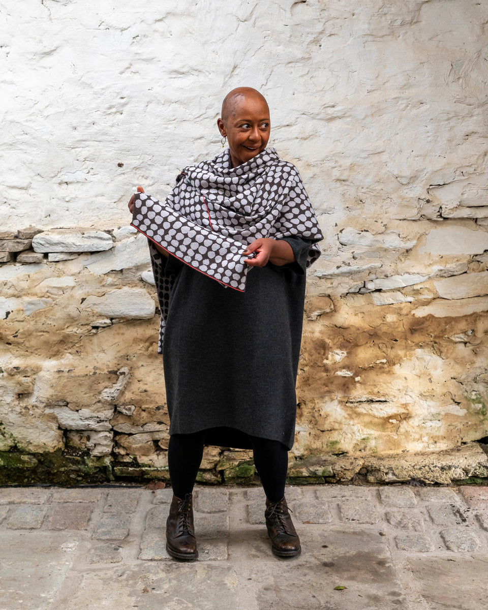 A black woman with a shaved head stands in a rustic stone building with whitewashed walls and a stone flat floor in Shetland. She wears a finely knitted modern shawl around her shoulders. It is charcoal grey with pale grey irregular dots and a thin meandering line in cherry red. She also wears a charcoal woollen dress in a loose fit, black tights and dark brown brogue boots.