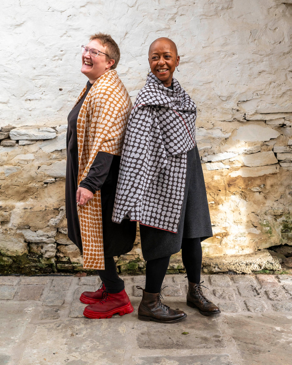 Two women stands in a whitewashed stone shed with flagstoned floor. On the left a white woman with fair hair wears a modern shawl in an ochre colour with irregular dots over a black tunic and leggings. On the right, a black woman with a shaved head wears the same shawl but in charcoal grey and pale grey over a grey woollen dress.
