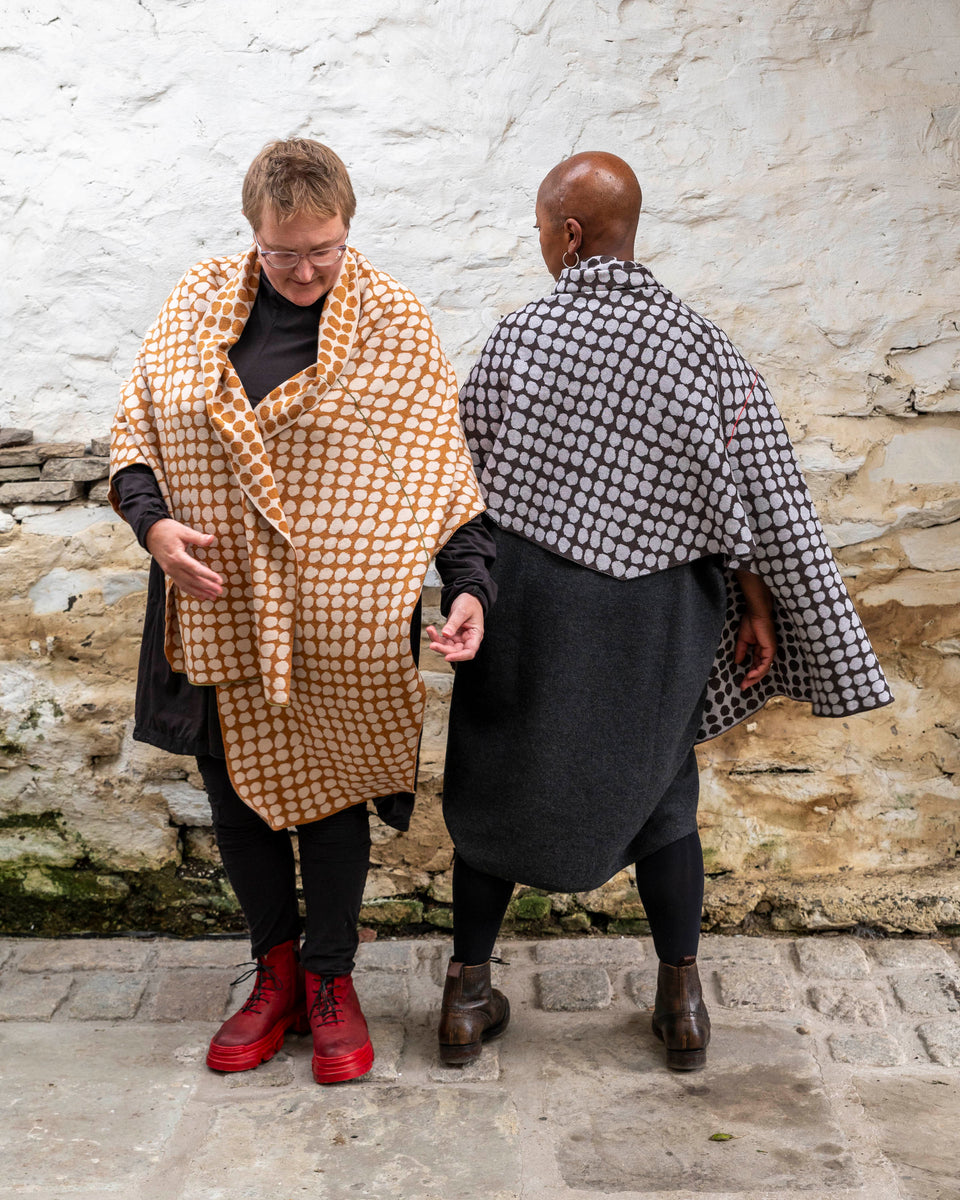 Two women wearing finely knitted shawls in an irregular dot pattern with contrast colour in a meandering line. On the left, a white woman with fair hair wears a shawl in ochre and cream, on the right a black woman with shaven head wears a shawl in charcoal and very pale grey