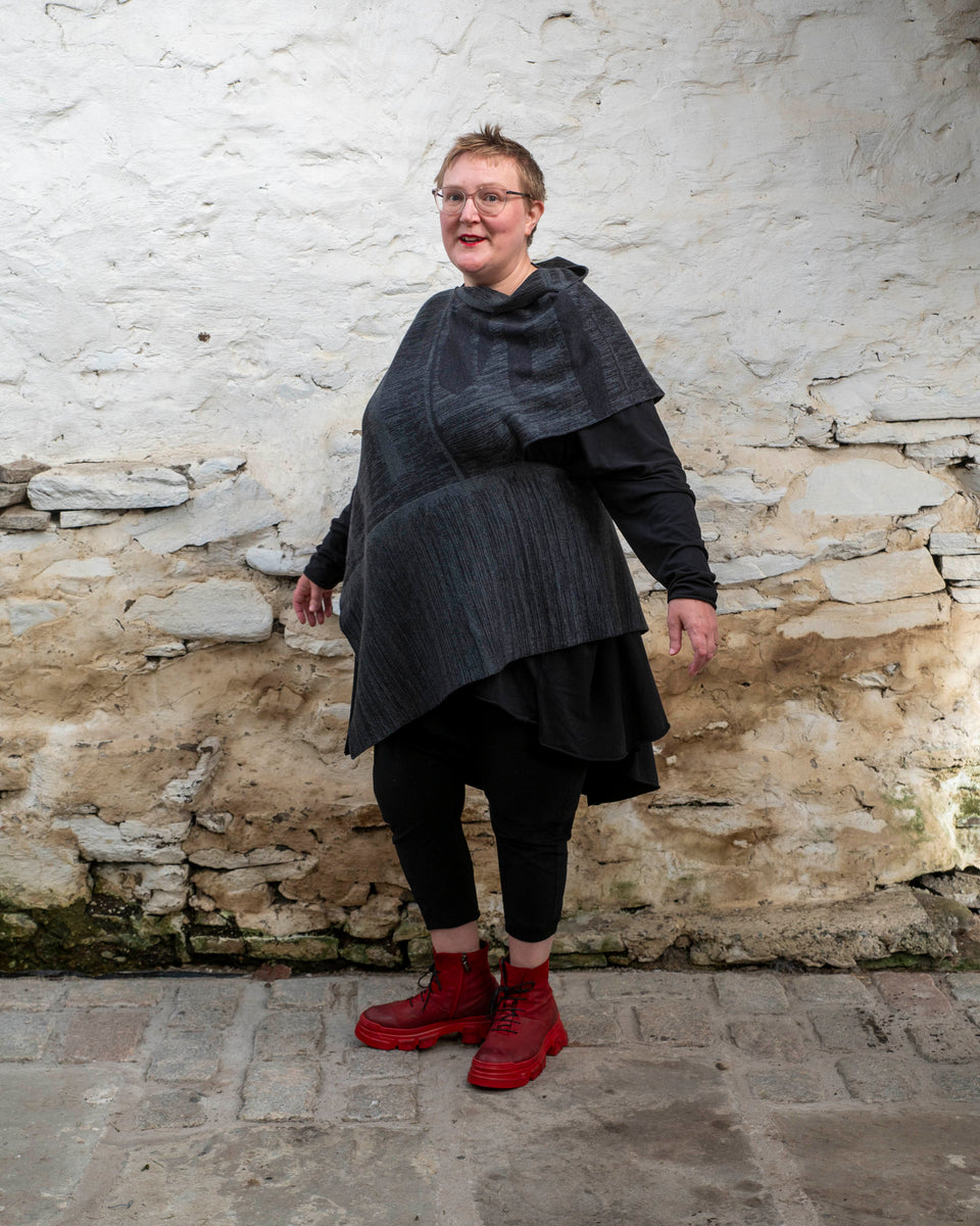 A white woman with short fair hair wears a long, asymmetric poncho with an abstract linear design in mid grey and charcoal over black leggings and a black tunic. She also wears chunky red leather boots.