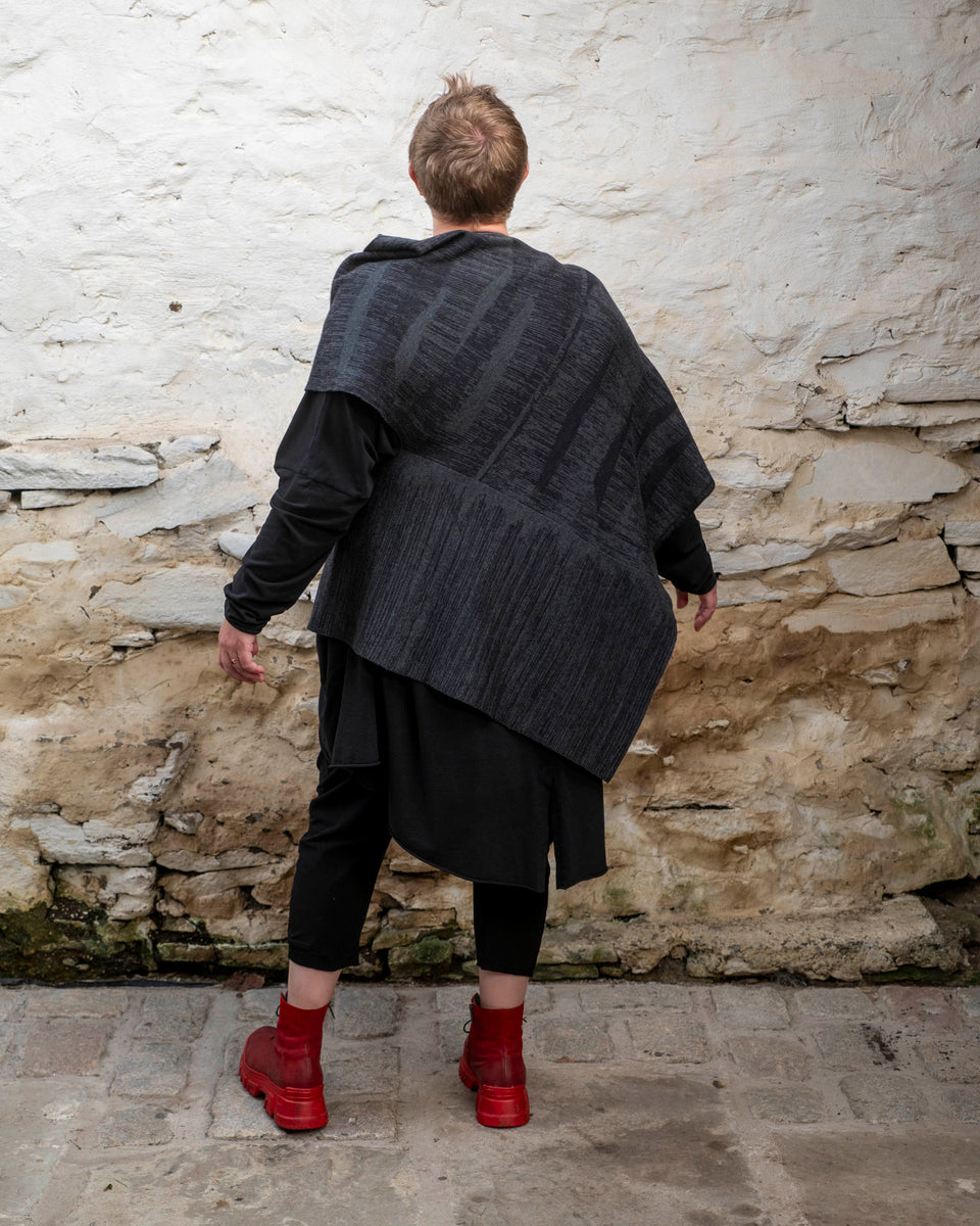 A white woman with short fair hair wears a long, asymmetric poncho with an abstract linear design in mid grey and charcoal over black leggings and a black tunic. She also wears chunky red leather boots.