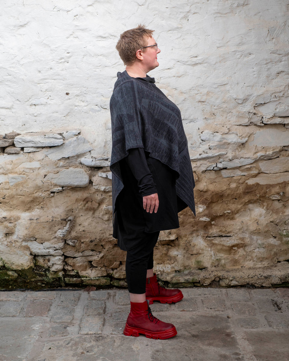 A white woman with short fair hair wears a long, asymmetric poncho with an abstract linear design in mid grey and charcoal over black leggings and a black tunic. She also wears chunky red leather boots.