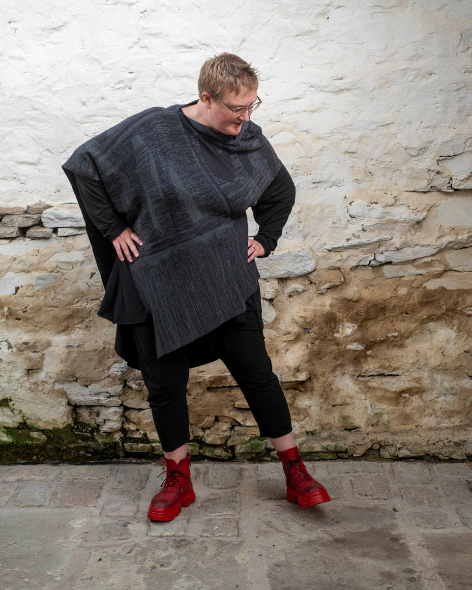 A white woman with short fair hair wears a long, asymmetric poncho with an abstract linear design in mid grey and charcoal over black leggings and a black tunic. She also wears chunky red leather boots.