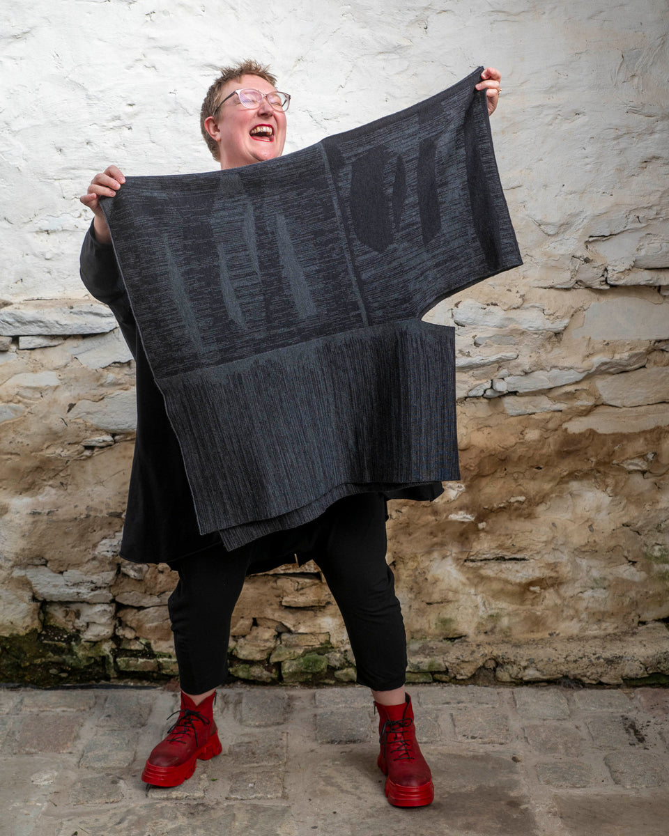 A white woman with short fair hair wears a long, asymmetric poncho with an abstract linear design in mid grey and charcoal over black leggings and a black tunic. She also wears chunky red leather boots.