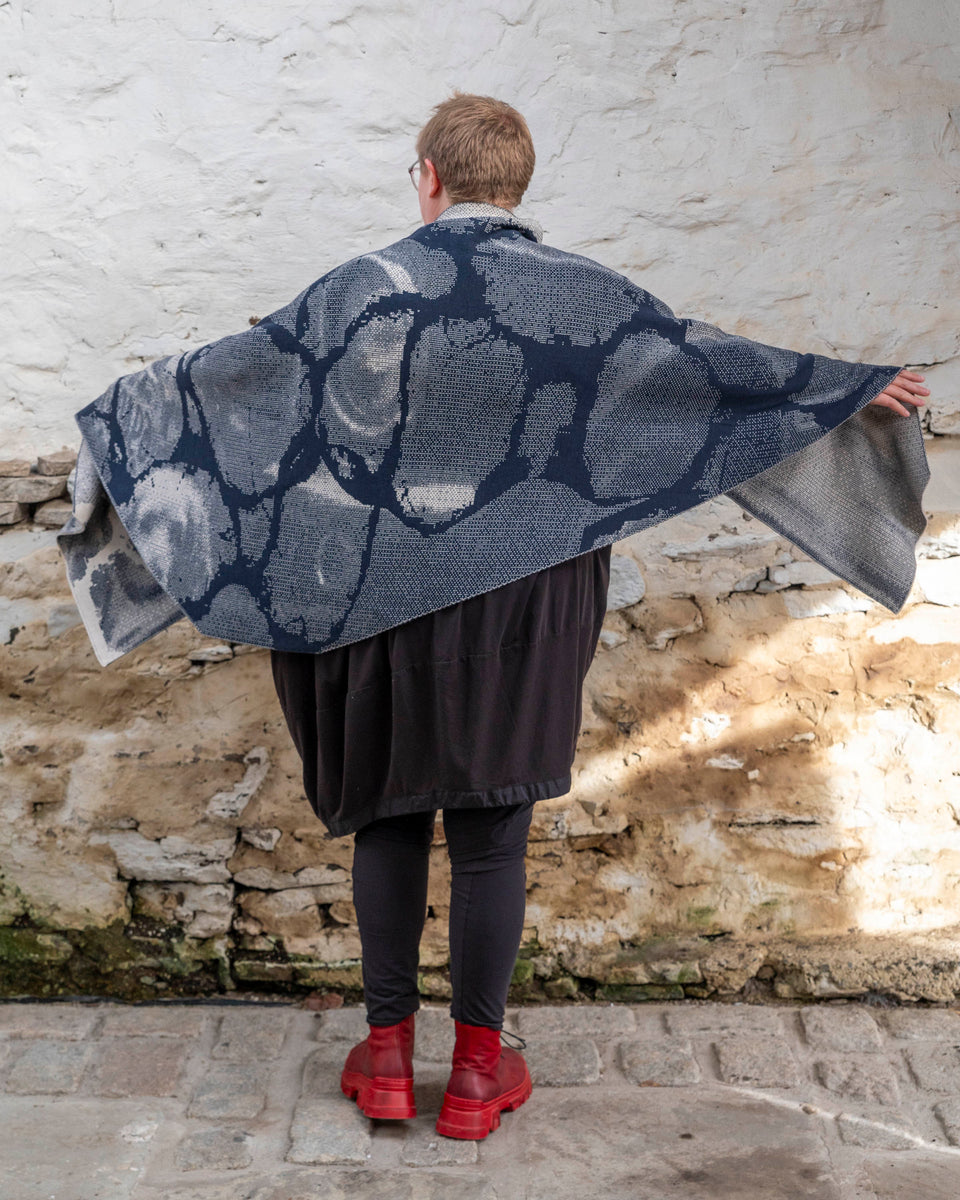 A white woman with short, fair hair stands inside a rustic stone building in Hoswick, Shetland. She wears an off-black tunic with matching leggings and red platform boots. She wears a navy and off white oversized shawl. She is standing with her back to the camera and holds out her arms to display the shawl