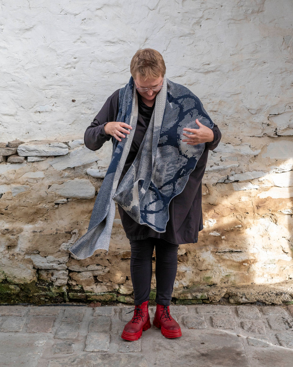 A white woman with short, fair hair stands inside a rustic stone building in Hoswick, Shetland. She wears an off-black tunic with matching leggings and red platform boots. She wears a navy and off white oversized shawl around her shoulders and looks down.