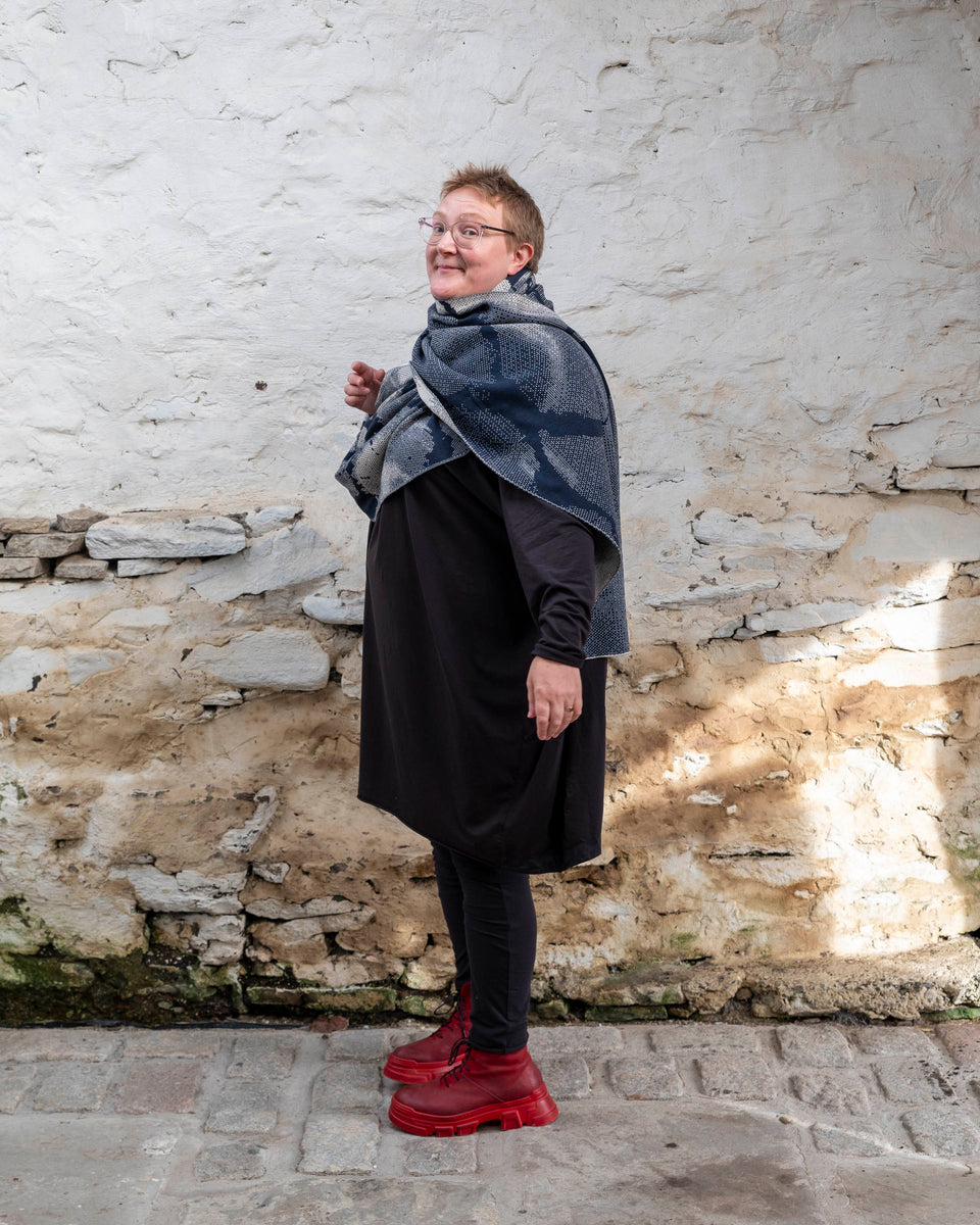 A white woman with short, fair hair stands inside a rustic stone building in Hoswick, Shetland. She wears an off-black tunic with matching leggings and red platform boots. She wears a navy and off white oversized shawl around her shoulders turns to look at the camera.