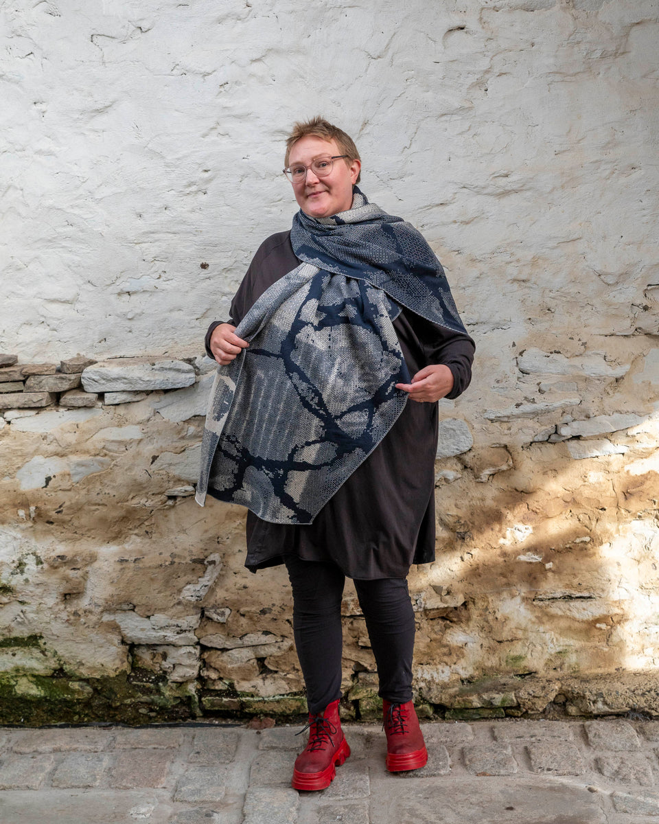A white woman with short, fair hair stands inside a rustic stone building in Hoswick, Shetland. She wears an off-black tunic with matching leggings and red platform boots. She wears a navy and off white oversized shawl which is arranged like a giant scarf, and holds of the front to show the pattern