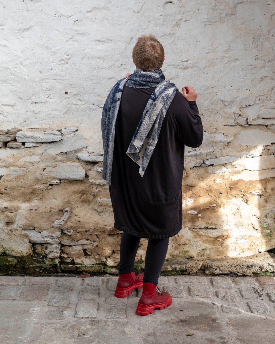 A white woman with short, fair hair stands inside a rustic stone building in Hoswick, Shetland. She wears an off-black tunic with matching leggings and red platform boots. She wears a navy and off white oversized shawl around her shoulders has her back to the camera