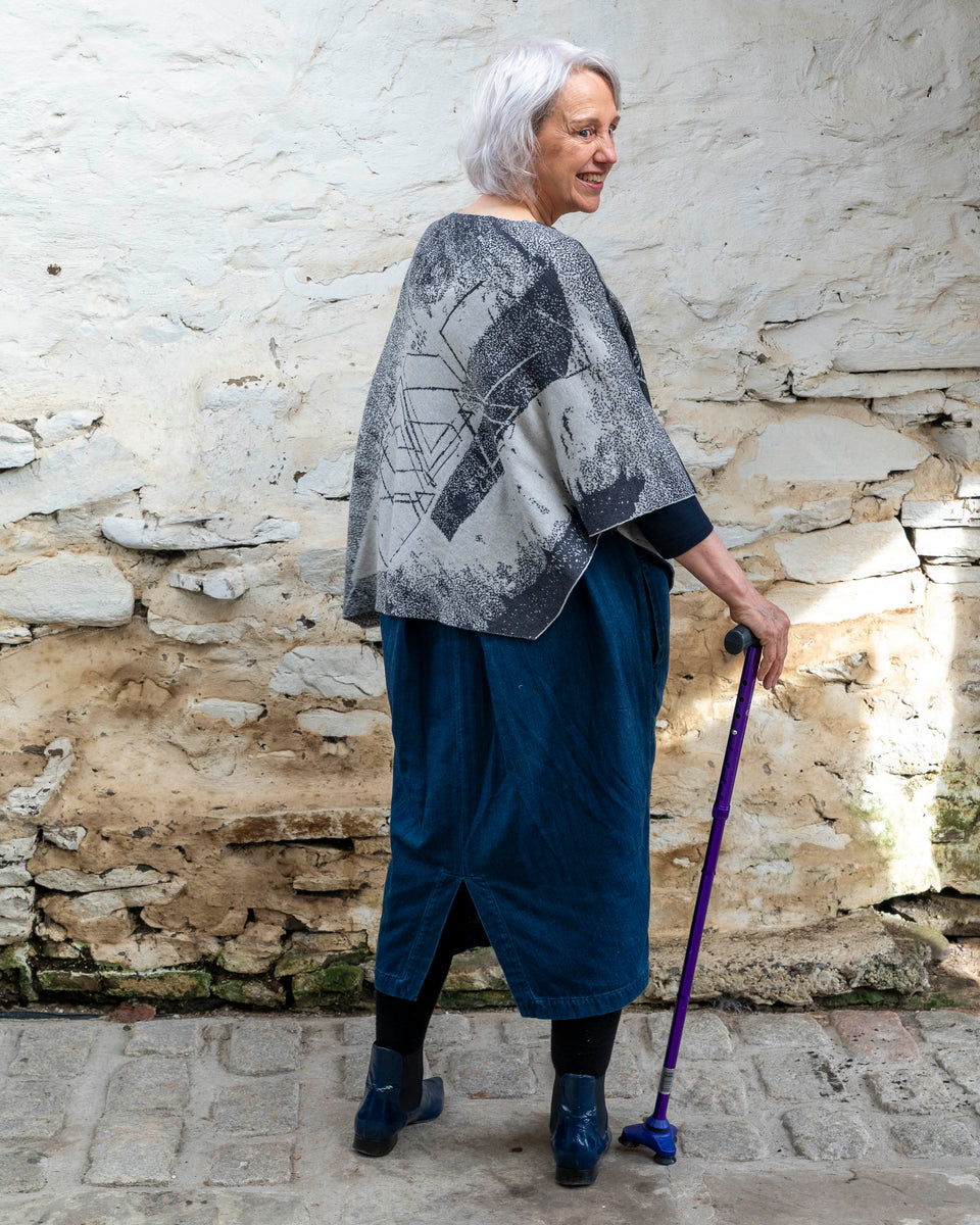 A woman with jaw length silver grey hair stands in a rustic, whitewashed stone building in Hoswick, Shetland. She holds a purple walking stick with her right hand and her back is to the camera, she turns her head over her right shoulder and smiles . She is wearing a finely knitted piece of contemporary Scottish knitwear - a cape in charcoal and soft white. underneath she wears a navy t-shirt and a loose indigo denim dress. She wears patent navy pointy dealer boots