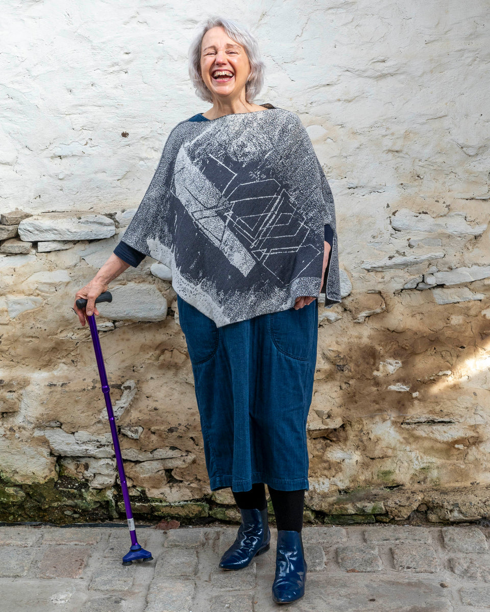 A woman with jaw length silver grey hair stands in a rustic, whitewashed stone building in Hoswick, Shetland. She holds a purple walking stick with  her right hand and laughs, thowing her head back. She is wearing a finely knitted piece of contemporary Scottish knitwear - a cape in charcoal and soft white. underneath she wears a navy t-shirt and a loose indigo denim dress. She wears patent navy pointy dealer boots.