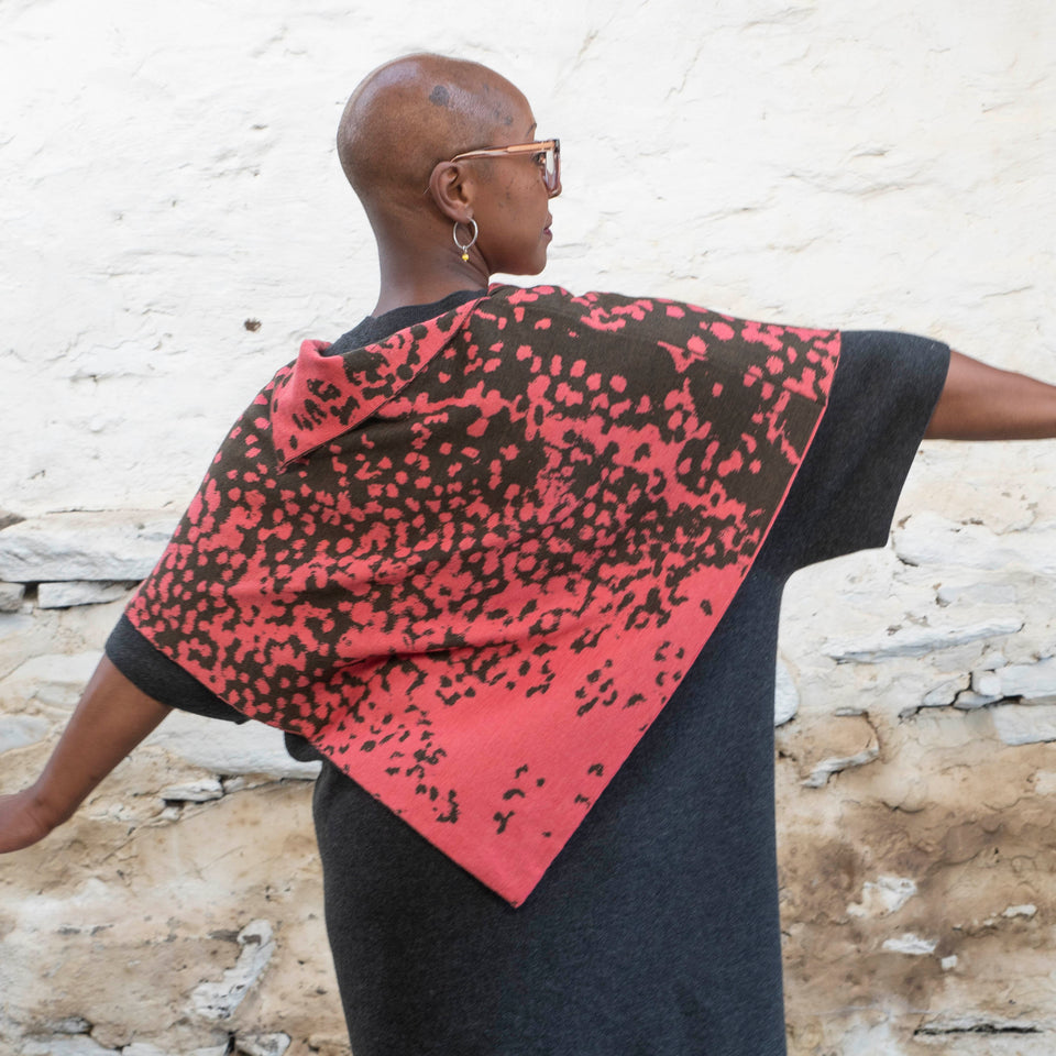 A black woman with a shaved wears tortoiseshell glasses and hooped silver earrings. She has her back to the camera and stretches out her arms. She is wearing a knitted cape with an abstract speckled pattern in pink and dark green over a charcoal grey dress. She's standing in a rustic, stone, whitewashed Shetland shed.