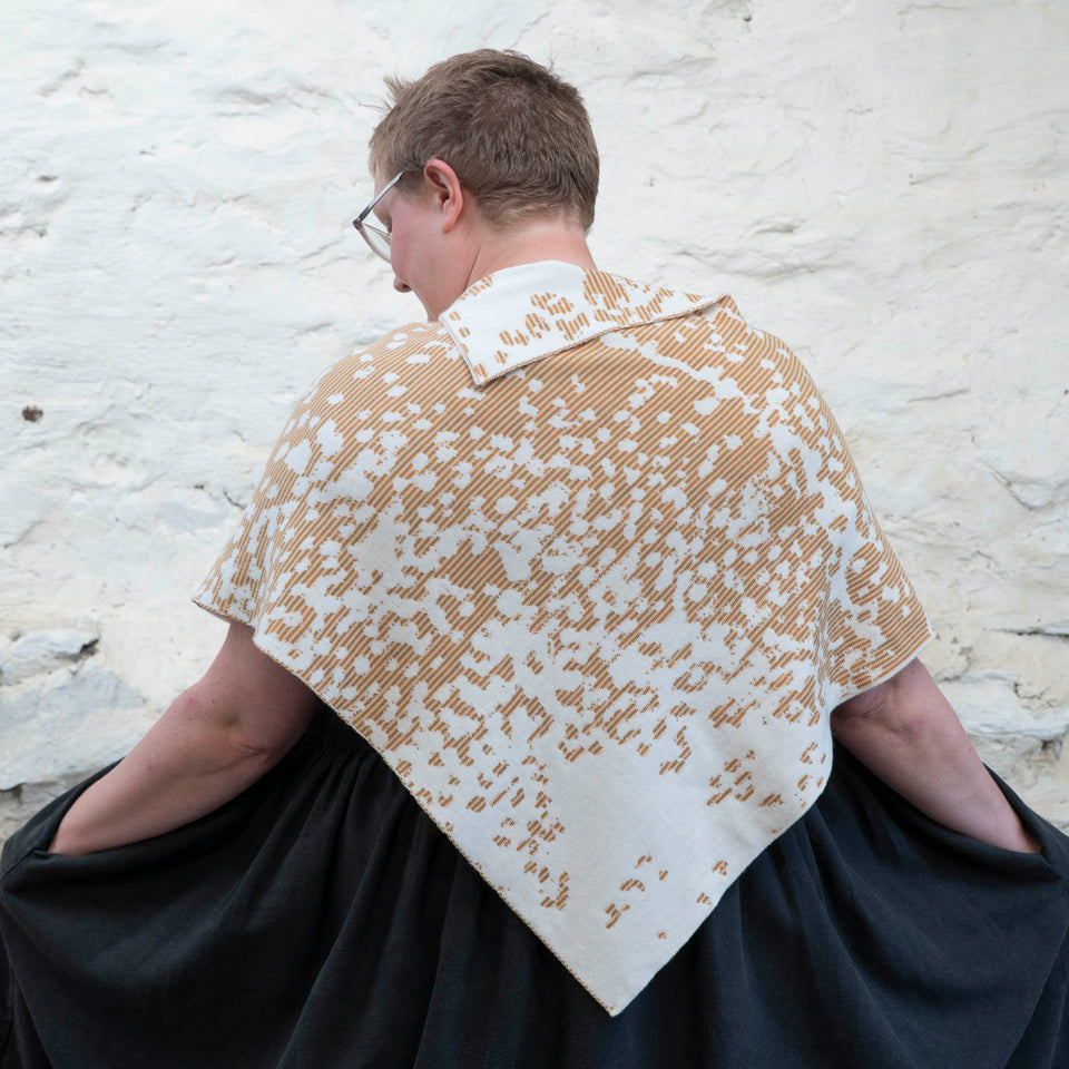 Felix Ford stands in a rustic, whitewashed stone shed in Hoswick, Shetland. A white woman with short, fair hair wears spectacles and - over a black linen dress with gathered skirt - a finely knitted cape in a speckled pattern in cream, gold and brown. She has her back to the camera and has her hands in the pockets of her dress.